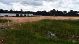 Cleared construction site with grass, dirt, traffic cones, equipment and a long metal building under a cloudy sky.