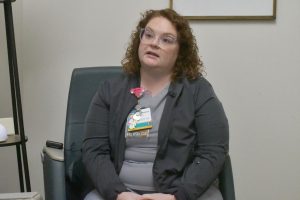 Marissa Black sits in a chair during an interview in an office. She is wearing gray scrubs and a dark jacket and has an employee badge clipped to her shirt.