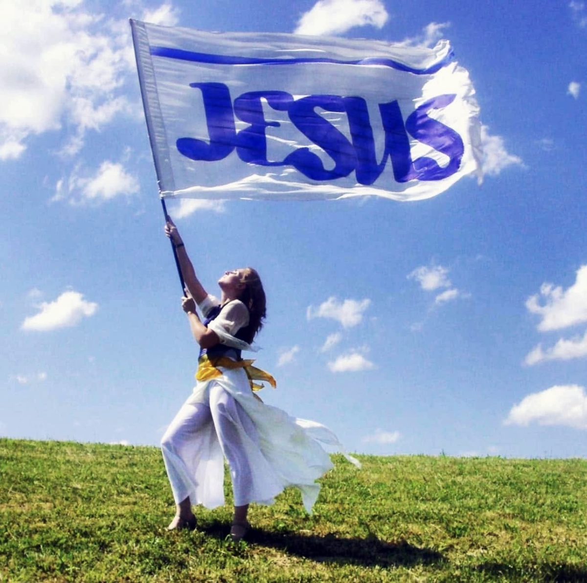 A woman stands barefoot on a grassy hill holding a large white flag with “JESUS” written in blue letters. She looks upward as the flag billows in the wind beneath a bright blue sky with scattered white clouds.