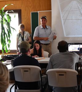 Dale Lucas stands at the front of a meeting room holding documents while presenting evidence of vandalism during a Longleaf Trace board meeting. Attendees are seated in the foreground, and security camera images are displayed on a projection screen.