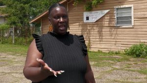 A woman wearing a black blouse gestures with one hand while standing outside a wood-sided building.