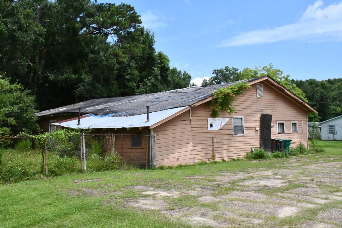 A wood-sided building in disrepair with damaged siding, patches of blue tarp on the roof and vines growing over the front roofline.
