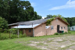 A wood-sided building in disrepair with damaged siding, patches of blue tarp on the roof and vines growing over the front roofline.