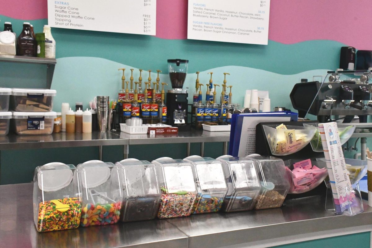 The service counter inside Bela's Coffee & Creamery in Diamondhead displays containers of colorful ice cream toppings in the foreground, with coffee syrups, a grinder, espresso machines and menu boards behind the counter.