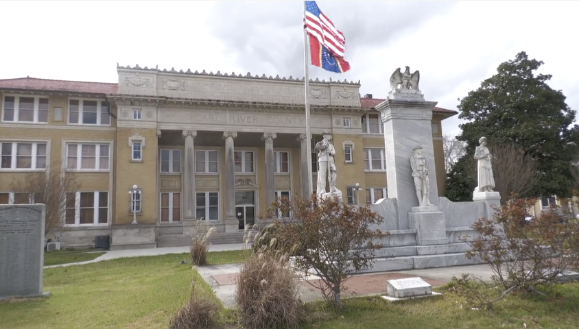 The Pearl River County Courthouse, a light-colored concrete government building in Poplarville, is shown behind a lawn with the U.S. and Mississippi flags, monuments, and landscaped walkways.