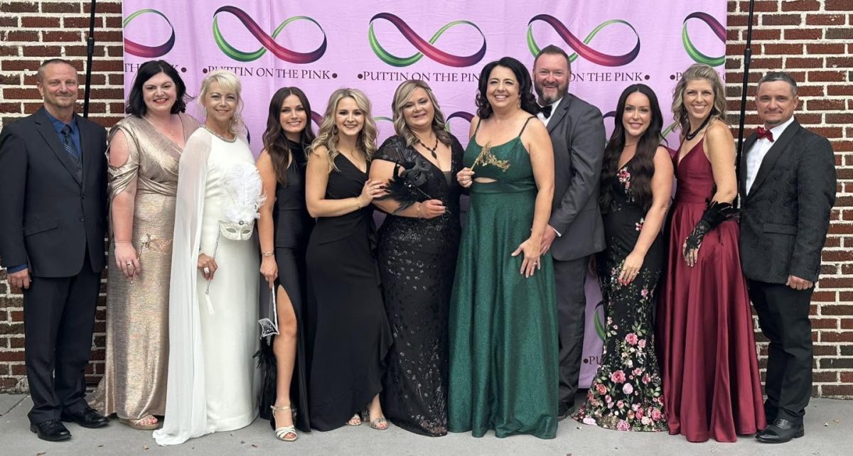 Eleven people dressed in formal gowns and suits pose in front of a pink Puttin' On The Pink backdrop set against a brick wall.