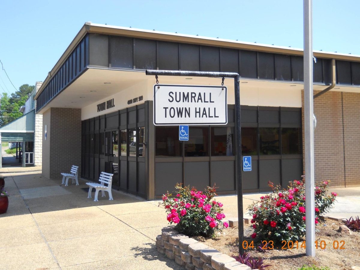 A white sign reading “SUMRALL TOWN HALL” stands in front of the Sumrall Town Hall building at 4880 Mississippi Highway 589, with benches, accessible parking signs and flower beds visible.