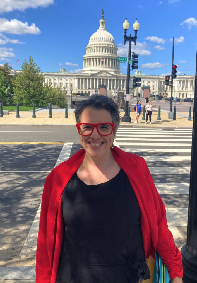 A woman wearing a red sweater, black shirt and glasses smiles near a crosswalk with a domed, Greco-Roman-style building in the background.