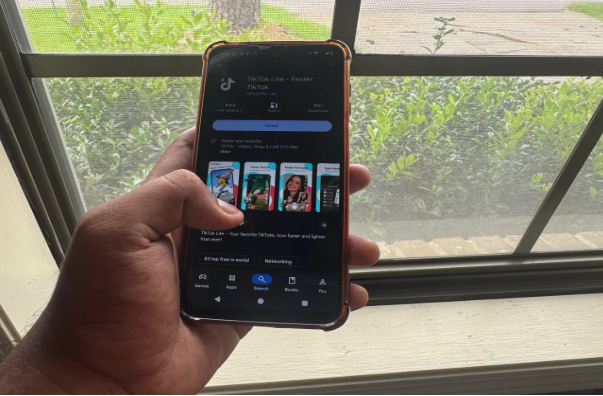 A person holds a smartphone displaying the TikTok Lite app page on the Google Play Store in front of a window with green shrubs outside.