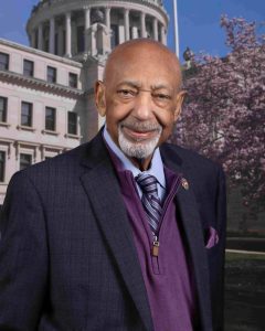 A man in a blue suit and tie poses for a headshot with a government building in the background.