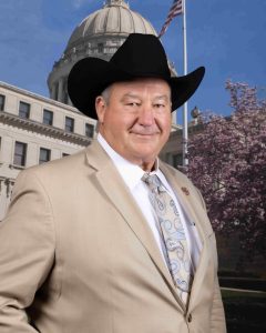 A man in a tan suit and tie poses for a headshot with a government building in the background.