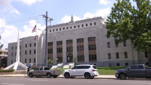 The Hinds County Courthouse stands behind a monument and a row of parked cars. The words “Hinds County Court House” are carved into the front of the building.